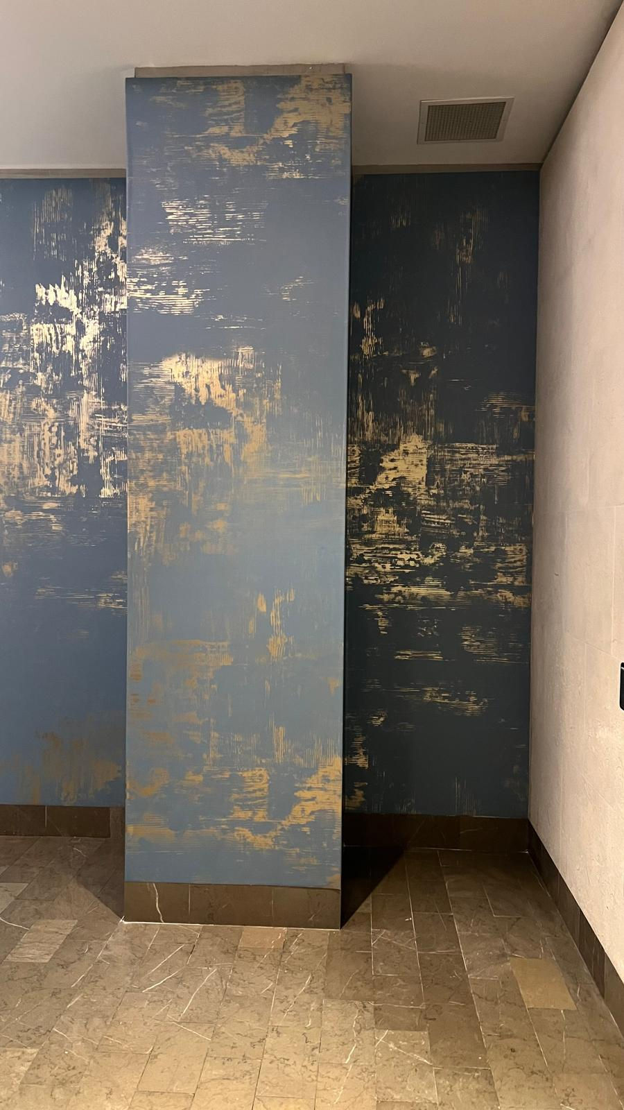 Textured blue and gold patterned wall in an interior setting, with taupe tile flooring. The scene is calm and modern. No text visible.