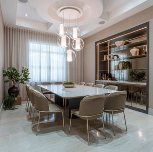 Elegant dining room with a marble table, beige chairs, and hanging glass lights. Shelves with decor and plants in the background; a calm ambiance.