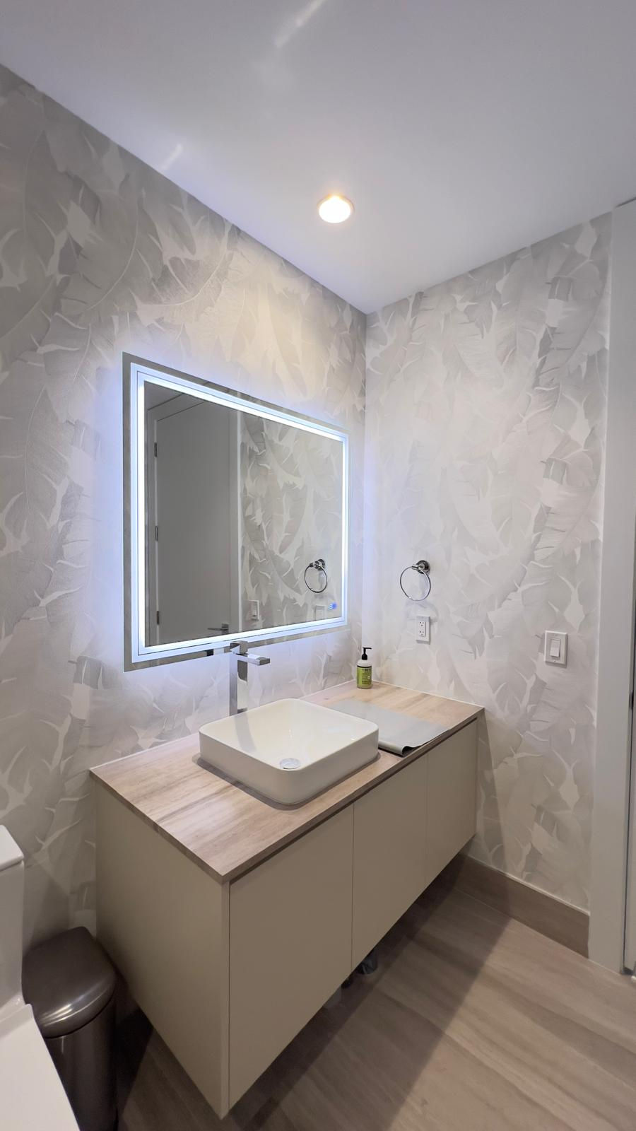 Modern bathroom with a lit mirror, beige vanity, and vessel sink. Leaf-patterned wall, wooden floor, and a soap dispenser create a clean look.