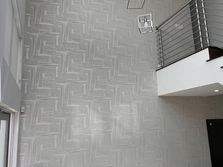 Soaring two-story entrance foyer wall at Oasis Park Square at Doral installed with GUH56738 Guimar Urbina Home greige geometric wallcovering behind suspended cube pendant lamps and a second-floor mezzanine.