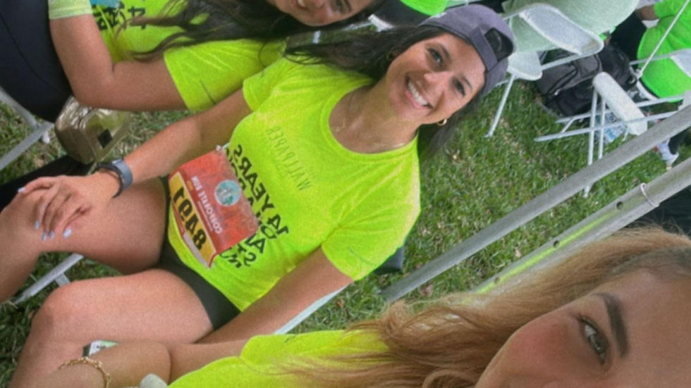 Women in neon green shirts, smiling and taking a selfie under a canopy. Others sit and stand nearby, creating a lively, outdoor event vibe.