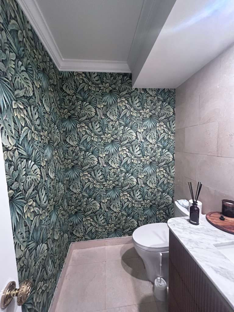 Bathroom with leafy green wallpaper, beige tiles, white marble counter, round mirror, scent diffuser, and a toilet. Calm, tropical vibe.