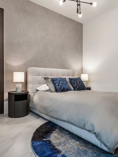 Modern bedroom with a grey upholstered bed, blue pillows, round two-tone rug, dark wood doors, and white bedside lamps, creating a cozy feel. Located at Lotus Boca Raton