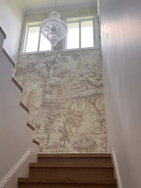 Two-story staircase accent wall installed with a custom wall mural in Miami, featuring a sepia-toned etched forest landscape beneath two sunlit windows and a crystal chandelier.