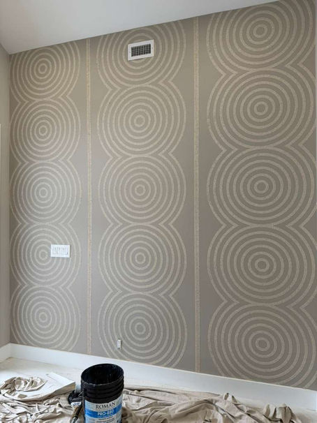Gray wall with concentric circle patterns, paint bucket labeled "Roman PRO-880" on a tarp. Minimalist room with a vent and light switches.