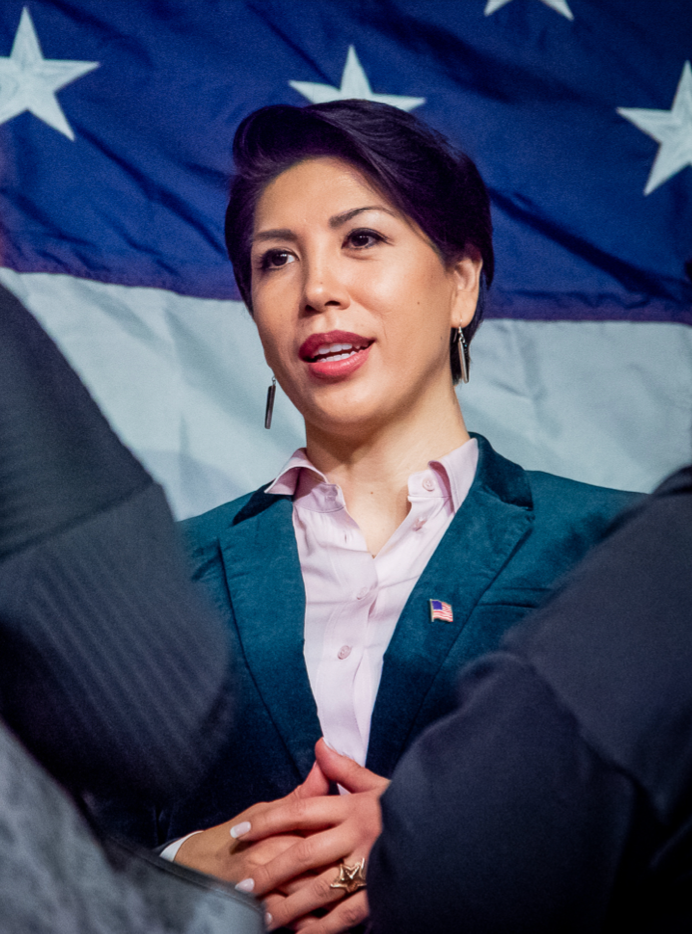 Portrait photograph of woman in front of the American flag. She has light brown skin, short dark hair, and is slightly smiling. She is wearing a dark blue blazer with a small American flag pin and a light pink button up shirt.