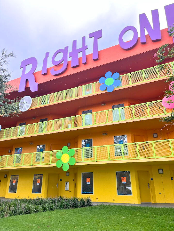 Colorful building with "Right On" in purple letters. Yellow balconies, vibrant flowers, orange facade, and green grass. Playful mood. The 1960's section at Pop Century is currently undergoing room refurbishment.