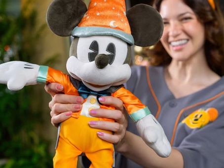 A woman smiles, holding a Mickey Mouse plush in orange wizard attire. She wears Mickey-themed Halloween headband and shirt in cheerful setting.