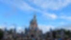 A pink castle with blue spires stands under a bright blue sky. Crowds of people walk nearby, creating a lively atmosphere.