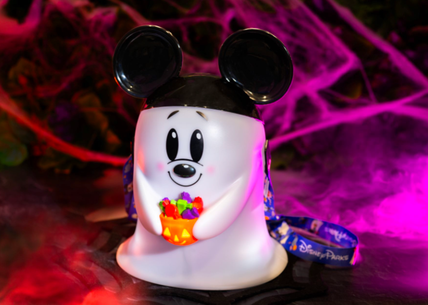 Ghost-shaped container with mouse ears holds a candy bowl. Background has pink and purple lighting with cobwebs. Disney Parks lanyard visible.