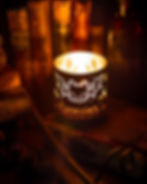 Lit candle with bat design sits on a table, surrounded by old books. Text reads "Gideon's Bakehouse." Warm, cozy ambiance.