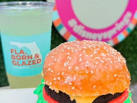 Cup and donut resembling a burger, on white table. Green drink in clear cup labeled "FLA. BORN & GLAZED." Background: colorful sign.