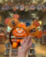Hands hold Mickey-shaped pumpkin headband in a decorated store with Halloween sign, orange pumpkins, and festive merchandise visible.