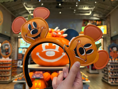 Hands hold Mickey-shaped pumpkin headband in a decorated store with Halloween sign, orange pumpkins, and festive merchandise visible.