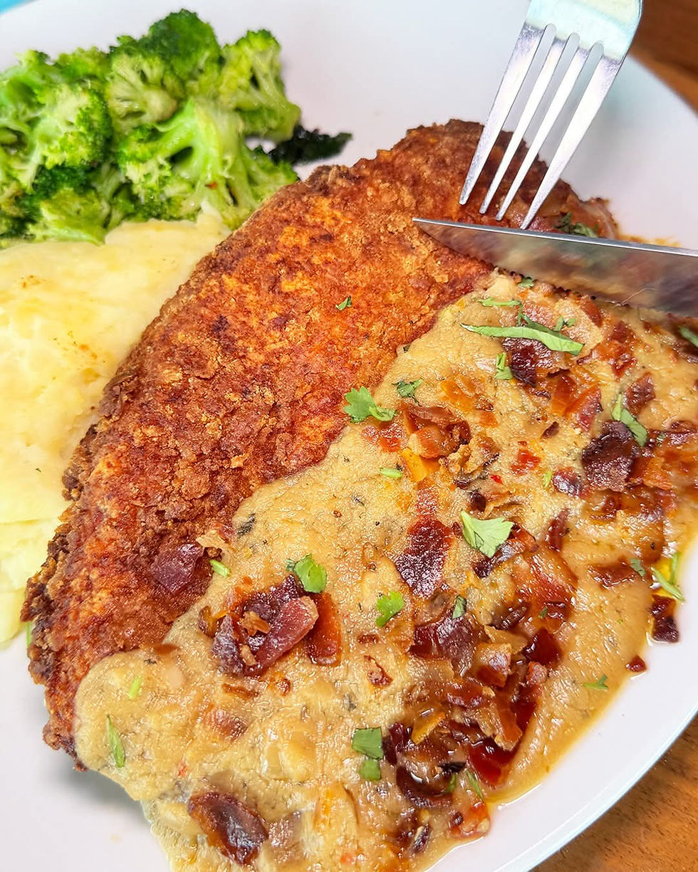 Close-up of a plate with mashed potatoes, broccoli, and a breaded steak topped with gravy and herbs. A fork and knife cut the steak.