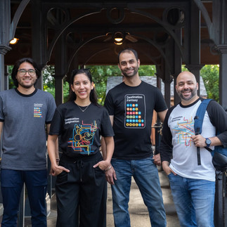 Four adults stand together smiling under a covered walkway at a Disney park, each wearing themed T-shirts from the Passport Collection. The shirts feature colorful, metro-inspired graphics with Disney park references, including designs labeled “Destination Tomorrow,” “Destination Fantasy,” and “Destination Imagination.”