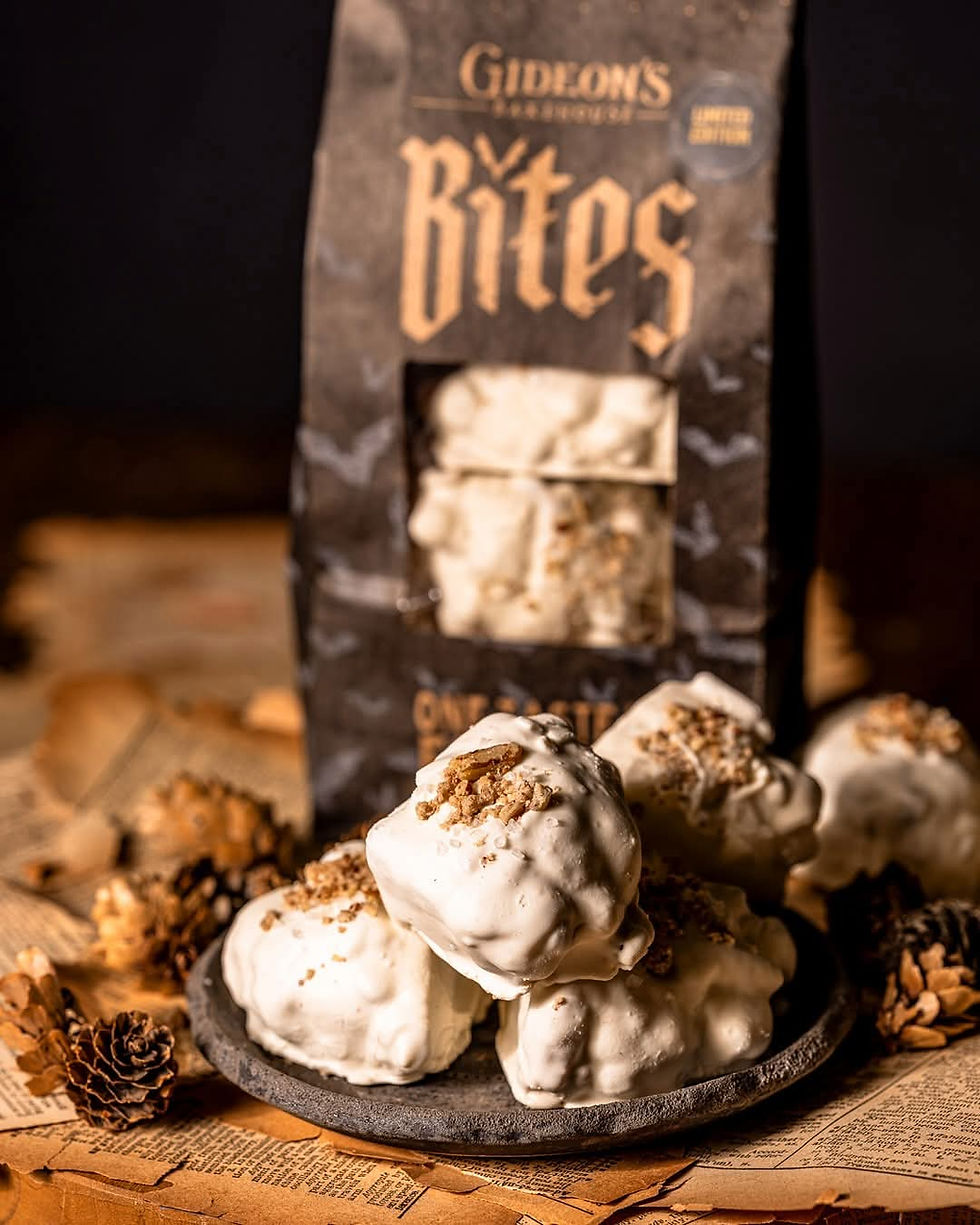 A plate of white chocolate-coated treats with crumble toppings is set on aged paper. Background shows "Gideon's Bites" on packaging. Cozy vibe.