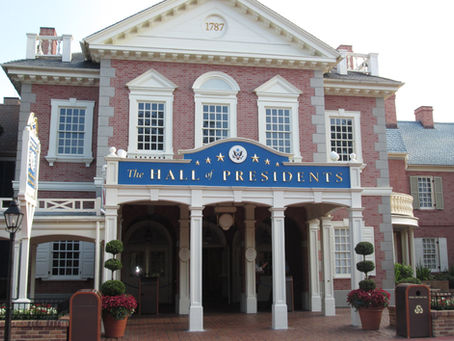 Red brick building labeled "The Hall of Presidents" with white columns, potted plants, and skylit windows. Sign reads "1787". Historic mood.