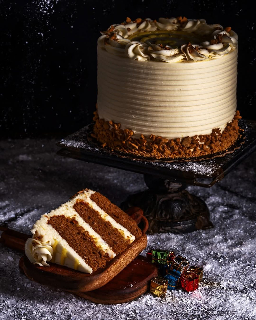A frosted cake with nut topping on a stand, a slice on a wooden board, surrounded by small colorful gift boxes on a snowy surface.