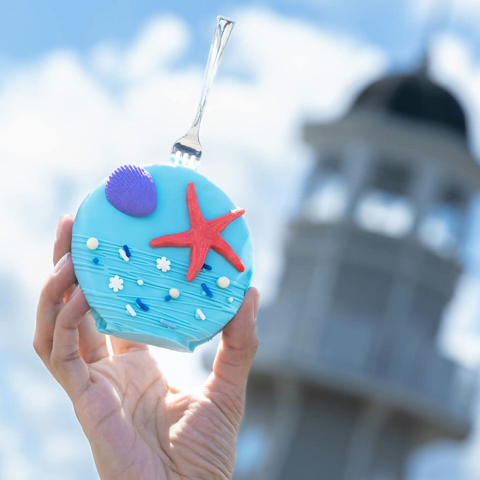 Hand holding a blue seashell-themed cookie with a red starfish, pierced by a fork. Blurred lighthouse and blue sky in background. This is the Little Mermaid cookie being offered at both the Yacht Club and Beach Club resorts.