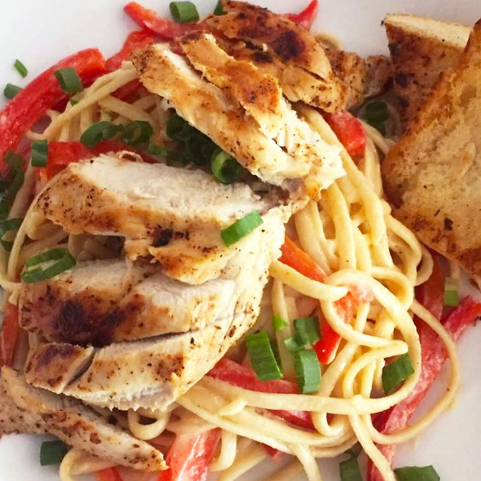 Grilled chicken on creamy pasta with red peppers and green onions, served with toasted bread in a white dish. Vibrant and appetizing.