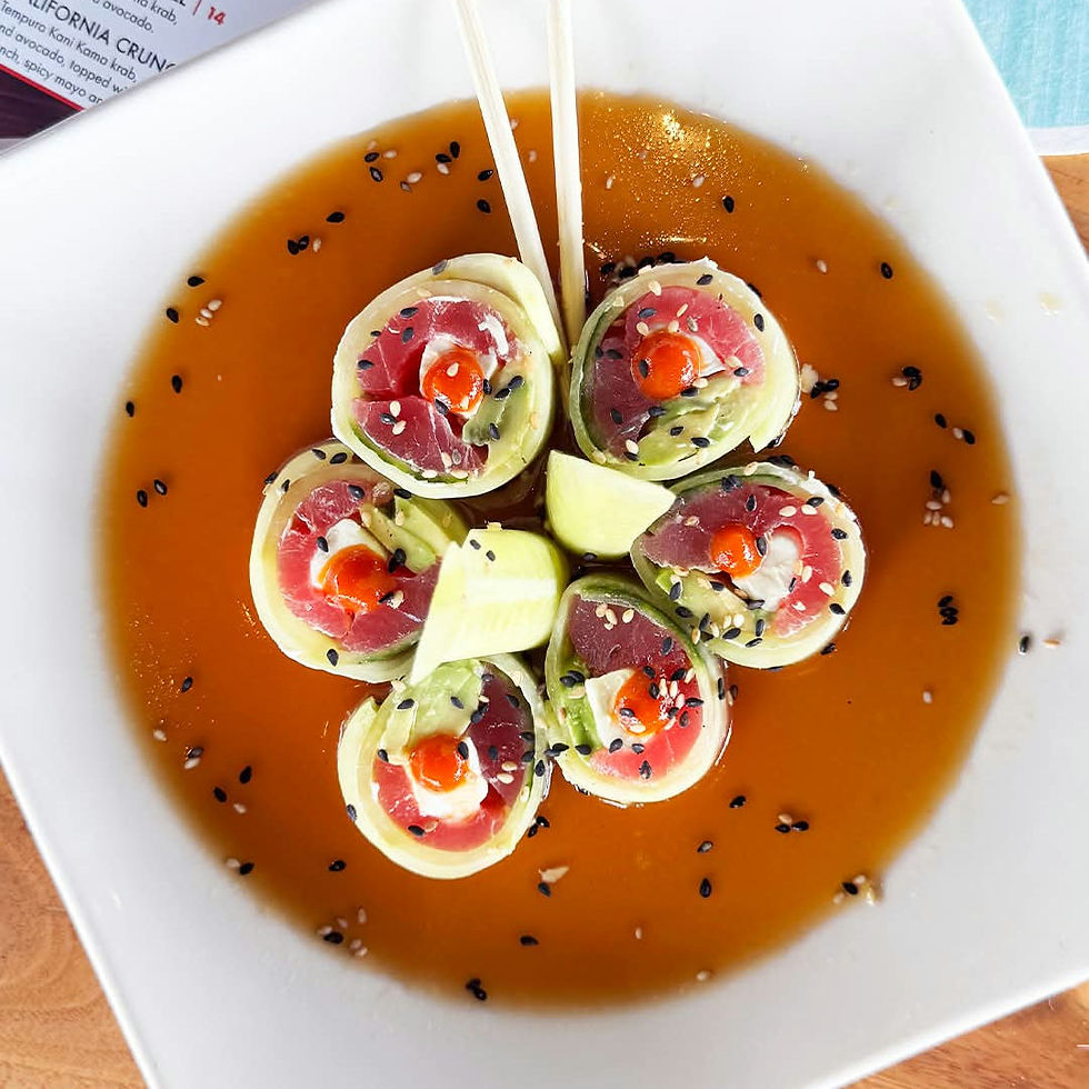 Sushi rolls topped with sesame seeds in a white dish with soy sauce, garnished with cucumber slices and chopsticks, on a wooden table.
