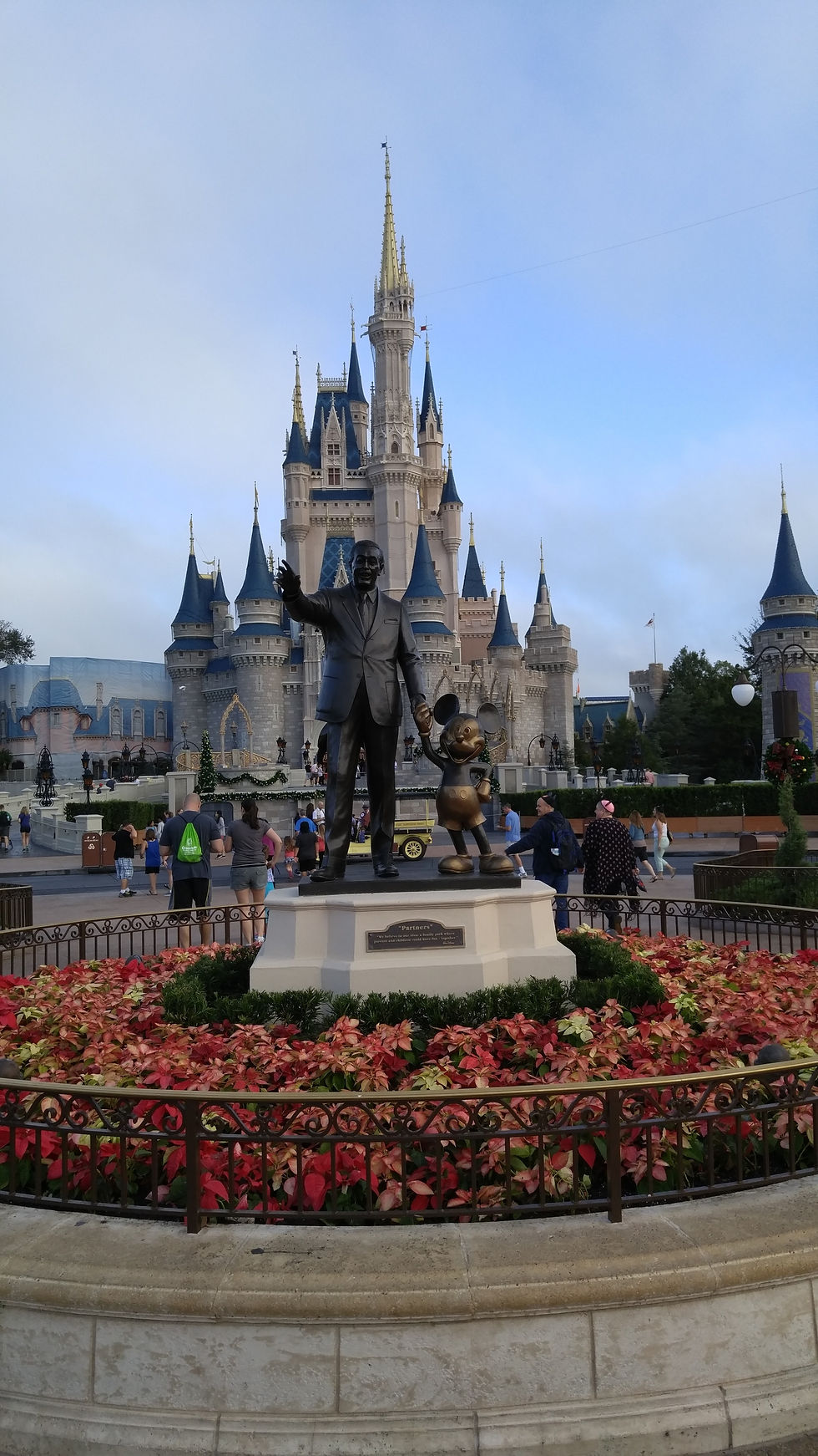 Statue of a man and cartoon character in front of a grand castle. People walk nearby, surrounded by vibrant red flowers and green plants.