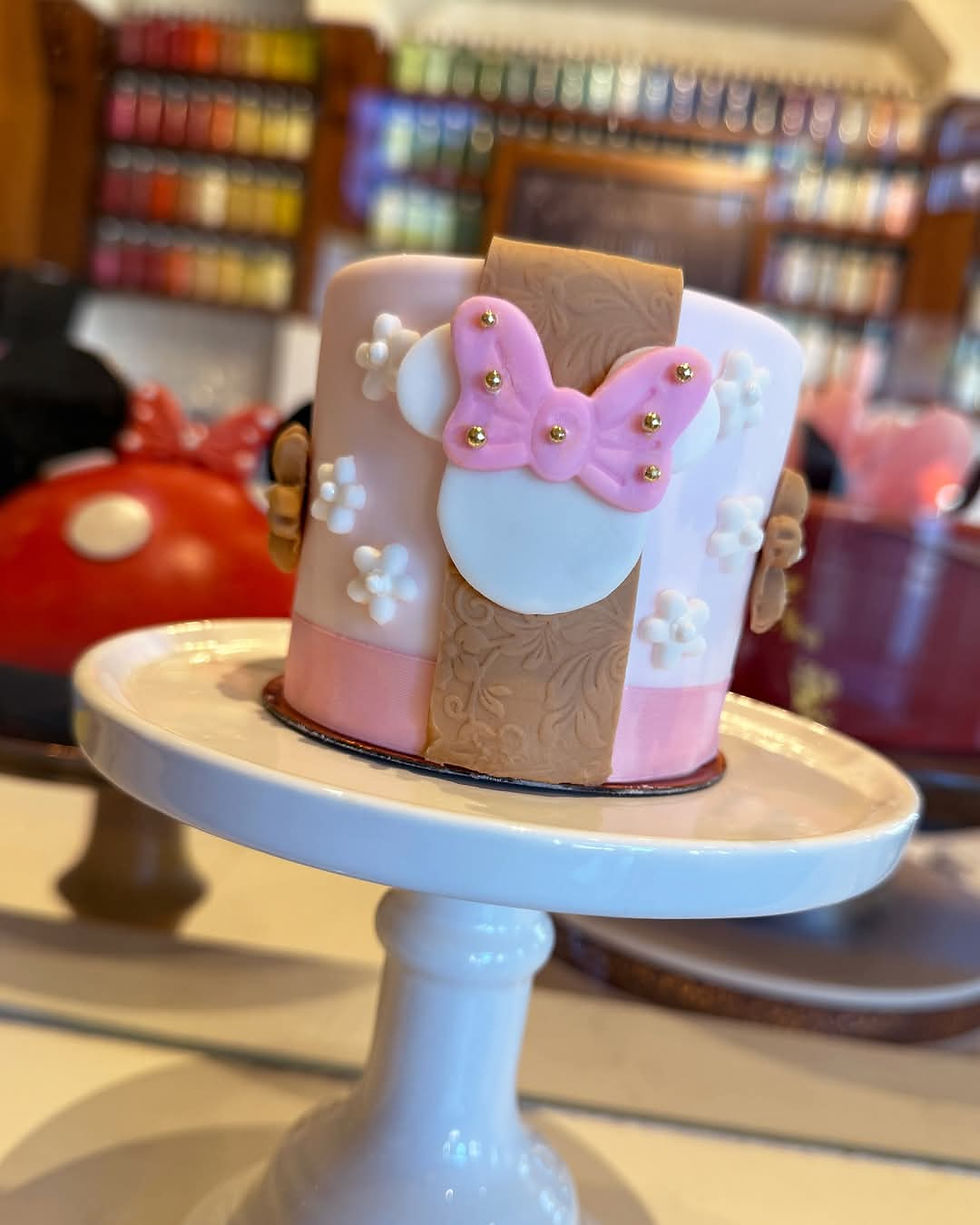 Pink and white cake on a stand, decorated with a pink bow and floral accents. Background shows colorful jars on shelves.