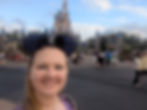 Woman in Mickey ears smiling in front of a blue castle. People and trees in the background. Sunny day with a festive atmosphere.