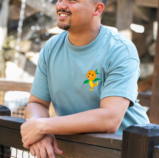 A smiling man leans against a wooden railing with a fountain blurred in the background. He is wearing a light blue T-shirt featuring an embroidered Orange Bird design on the chest.