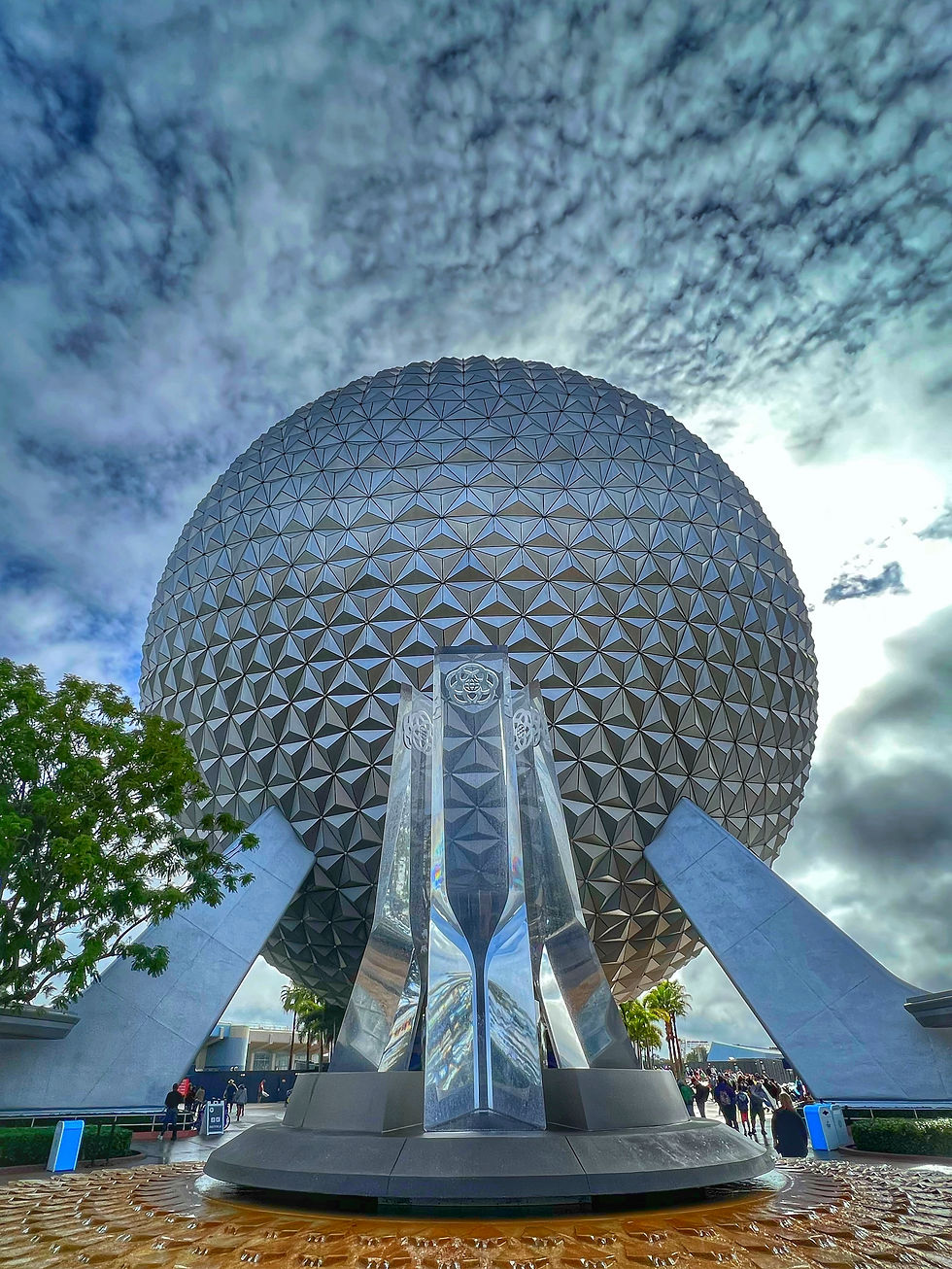 Geodesic sphere with reflecting monument in foreground, cloudy sky above. Crowds gather near palm trees, creating a lively atmosphere.