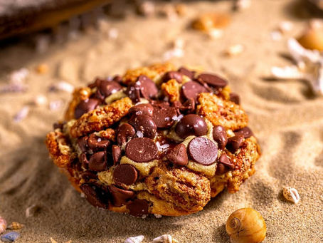 Chocolate chip cookie on sand with seashells and starfish around. Warm, inviting tones create a beach picnic vibe. The Sea Turtle Cookie is available at Gideon's until the end of July.