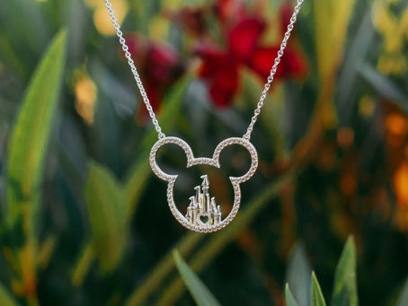 A Mickey Mouse-shaped necklace with a castle design hangs against a blurred green and red floral background, evoking a whimsical mood.