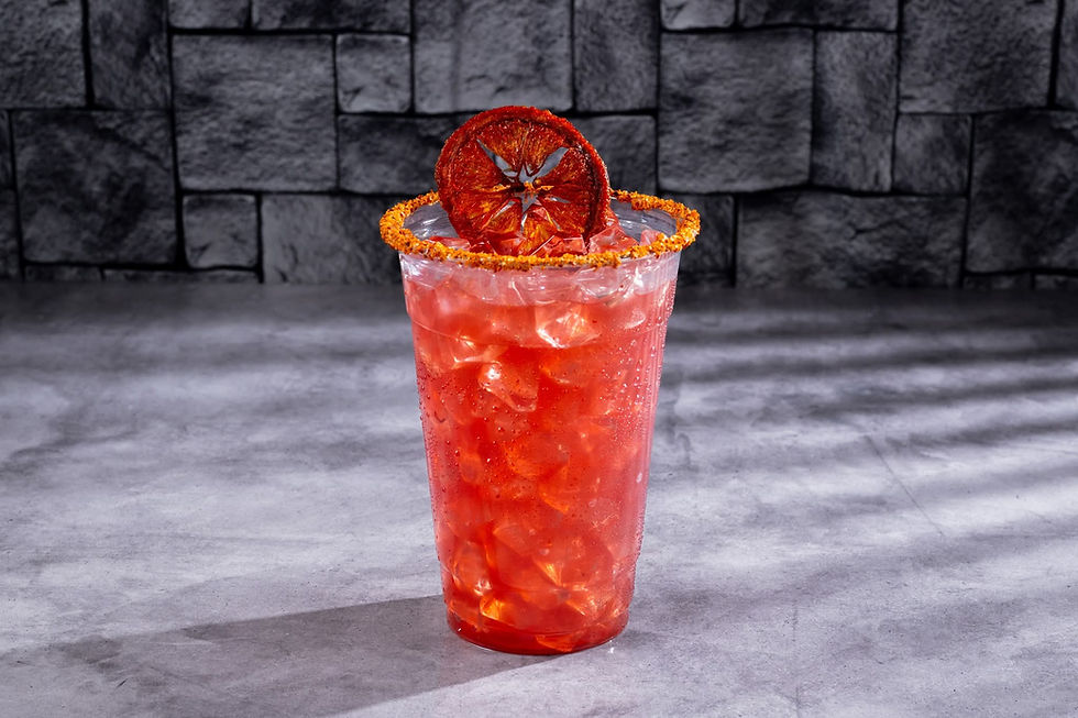 Red iced drink in a clear cup with chili rim, garnished with a dried orange slice. Set against a gray stone wall background.