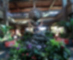 Wooden statue with a colorful lei stands on rocks in a tropical-themed lobby, surrounded by lush greenery and vibrant flowers.