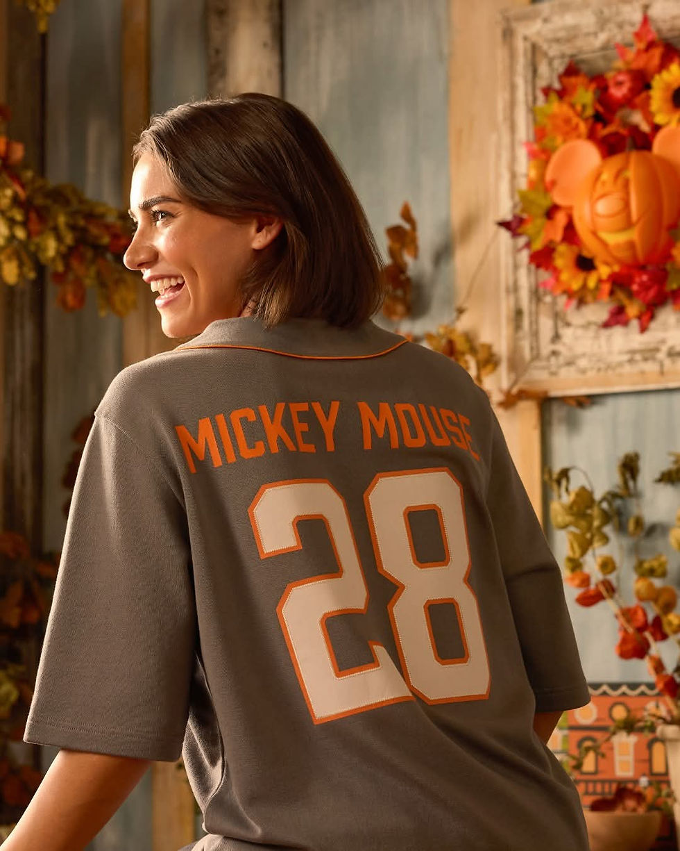 Woman smiling, wearing a gray "Mickey Mouse 28" shirt. Background has autumn leaves and a pumpkin-themed wreath on a rustic wall. Cozy mood.