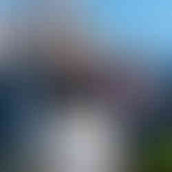 A guest stands in front of Sleeping Beauty Castle at Disneyland, wearing a cream-colored T-shirt from the Passport Collection. The shirt features colorful minimalist icons of Disney castles and attractions from around the world, with a bright sun design at the top. The castle is decorated with banners and a large “70” medallion for Disney’s 70th anniversary celebration.