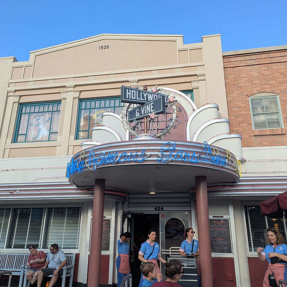 People outside Hollywood & Vine restaurant. Neon sign and art deco facade visible. Bright blue sky. Staff in blue shirts and pink aprons.