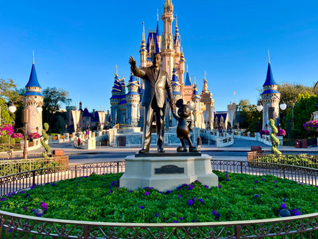 Statue of a man and a mouse character in front of a castle with blue spires, surrounded by green plants and purple flowers under a clear sky.
