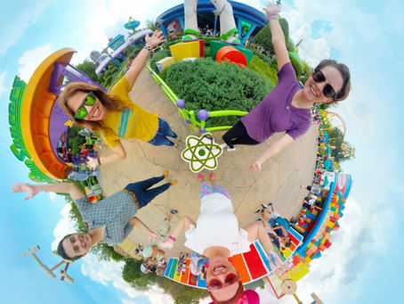 Four people pose excitedly under a giant Buzz Lightyear statue at a theme park. Bright colors and a "Pizza Planet" logo are visible.