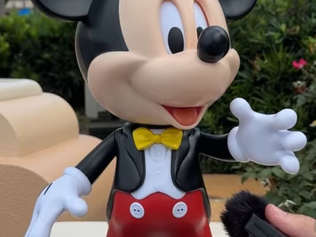 Toy figure of a famous cartoon mouse in red shorts and yellow bow tie. A hand holds a microphone near it. Background: trees and a building.