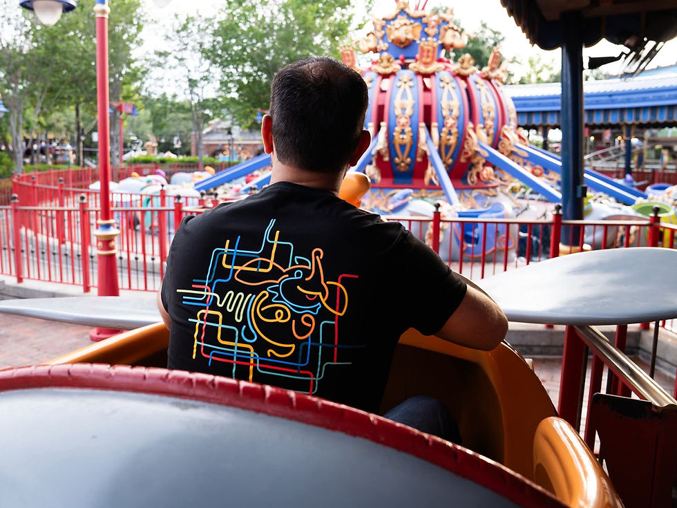 Man on a theme park ride facing colorful carousel, wearing a black shirt with abstract neon design. Trees and intricate ride details visible.