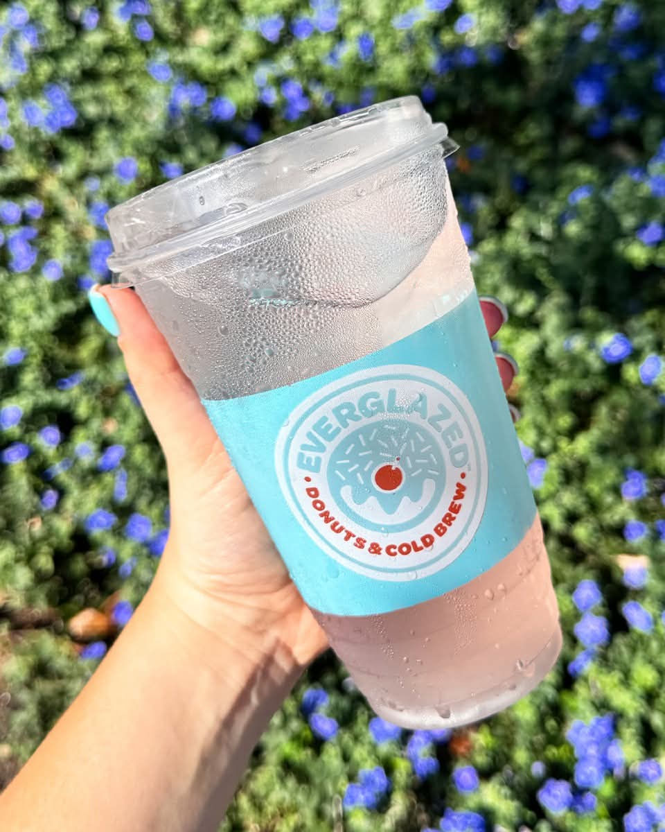 Hand holding a cold Everglazed drink against a backdrop of green grass and small purple flowers. Blue label with logo on the cup.