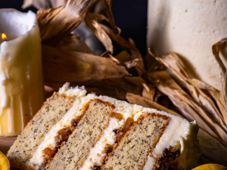 Slice of poppy seed cake with cream layers on a wooden board, surrounded by oranges, lemons, a lit candle, and corn husks. Warm tones.