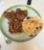 Plate with bear-shaped churros, a pecan cinnamon roll, and a blueberry pancake. Background is a light surface; utensils are visible.