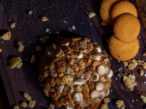 A top-down view of a large, thick cookie heavily topped with butterscotch chips, cookie crumbles, and sea salt. It rests on a dark wooden board engraved with the "Gideon’s Bakehouse" logo, accompanied by several round vanilla wafer cookies and golden crumbs.