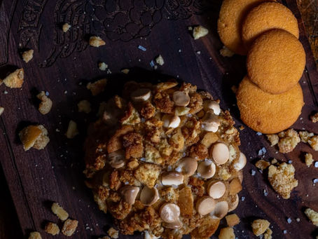 A top-down view of a large, thick cookie heavily topped with butterscotch chips, cookie crumbles, and sea salt. It rests on a dark wooden board engraved with the "Gideon’s Bakehouse" logo, accompanied by several round vanilla wafer cookies and golden crumbs.