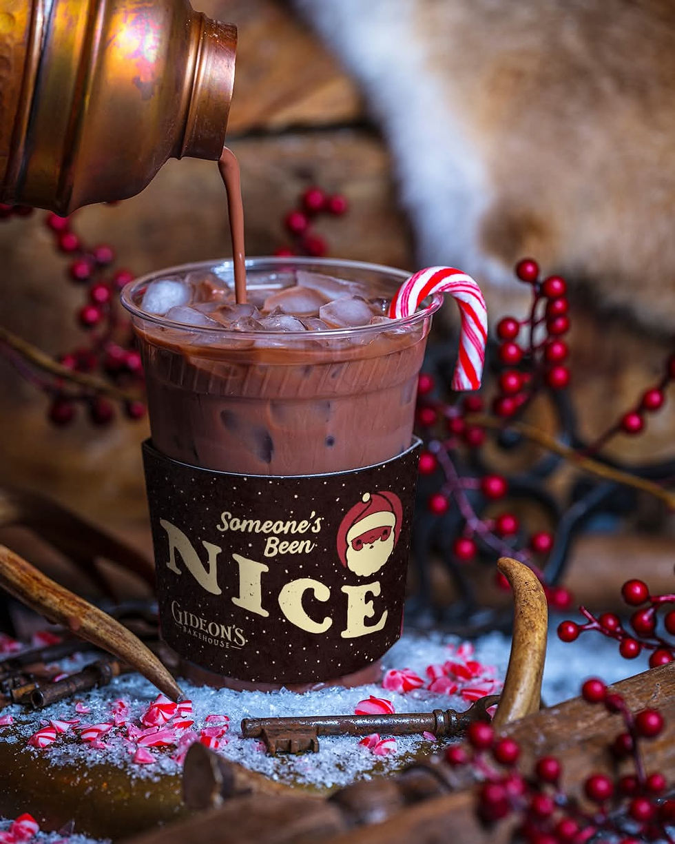 Iced chocolate drink in a cup labeled "Someone's Been Nice" with candy cane, placed among red berries and snow. Warm festive setting.