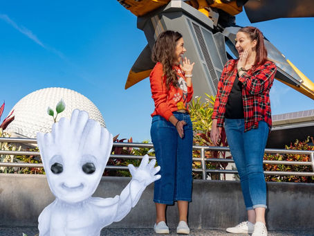 Two women wave at a small white Groot figure with a spaceship model and dome in the background, set against a clear blue sky.