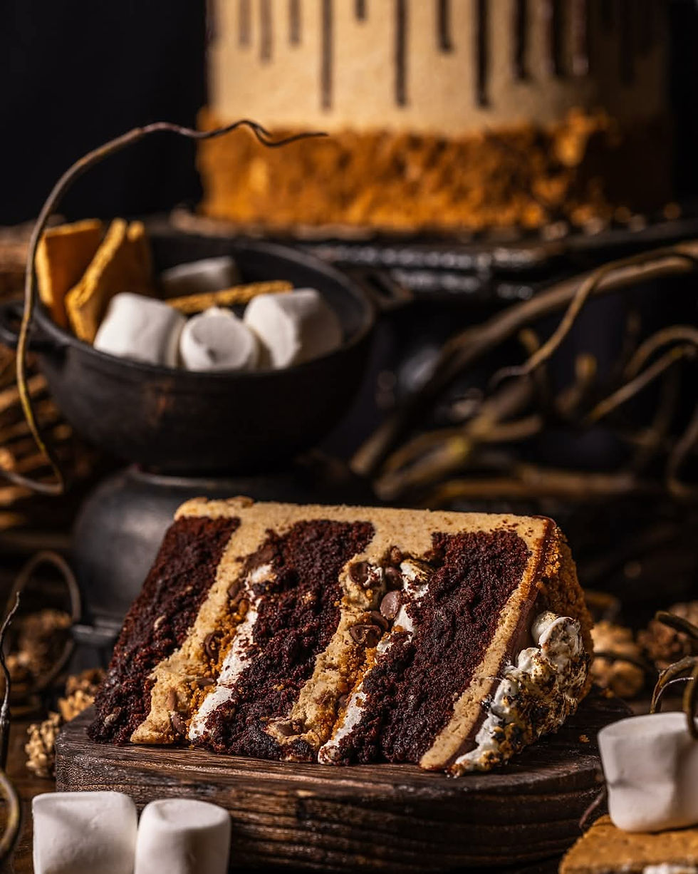 Slice of chocolate and marshmallow cake on a wooden board, surrounded by marshmallows and twigs, with a layered cake in the background. This is the S'mores cake being offered at Gideon's Bakehouse this week.